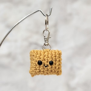 May include: A handmade, tan crochet keychain in the shape of a square with a smiling face. The keychain has a silver-coloured clasp and key ring. The crochet stitch creates a textured surface. The small, smiling face has two black bead eyes and a curved mouth.