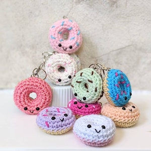 May include: A collection of eight crocheted donut keychains in various colors, including pink, green, blue, purple, and white. Each donut has a smiling face and colorful sprinkles.