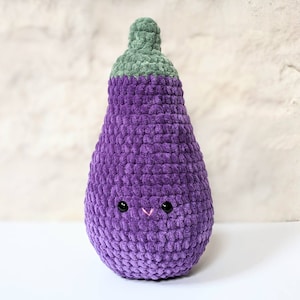 May include: A plush, purple eggplant with a green stem. The crocheted vegetable has a smiling face with black eyes and a pink stitched mouth. The soft toy is designed to look like a friendly eggplant.
