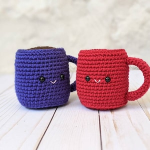 以下が含まれることがあります： 笑顔の顔をした、かぎ針編みで作ったコーヒーカップが2つ。1つは紫、もう1つは赤です。