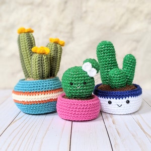 May include: Three crocheted cactus plants in colorful pots. The cacti are green with yellow flowers, and the pots are blue, pink, and white with smiling faces. A charming, handmade home decor item.
