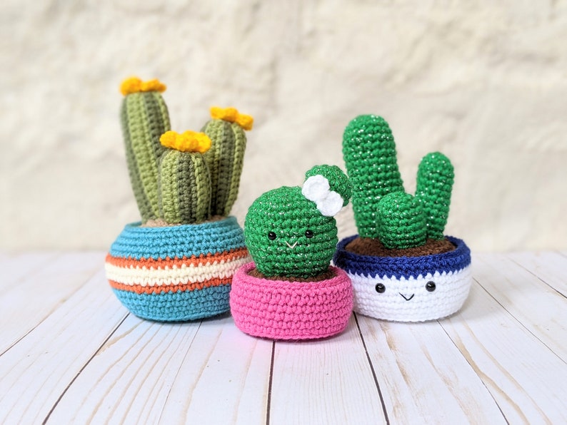 May include: Three crocheted cactus plants in colorful pots. The cacti are green with yellow flowers, and the pots are blue, pink, and white with smiling faces. A charming, handmade home decor item.