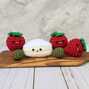 May include: Five crocheted amigurumi toys: a white brie cheese, two red strawberries with green leaves, and two green olives. The toys are arranged on a wooden cutting board.