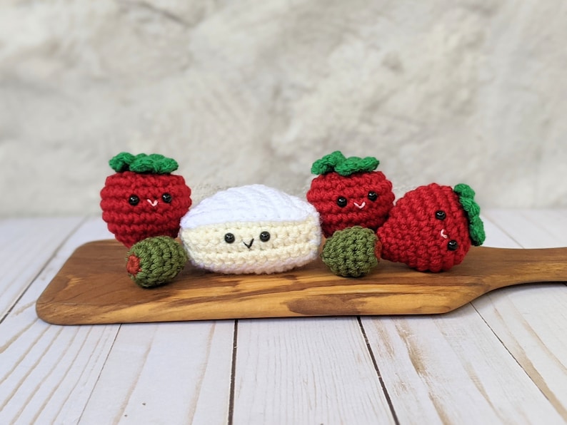 May include: Five crocheted amigurumi toys: a white brie cheese, two red strawberries with green leaves, and two green olives. The toys are arranged on a wooden cutting board.