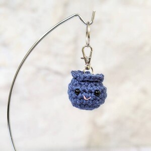 May include: A small, navy blue crocheted keychain shaped like a smiling blueberry. The blueberry has black bead eyes and a pink stitched mouth. It is attached to a silver-colored keychain clip.