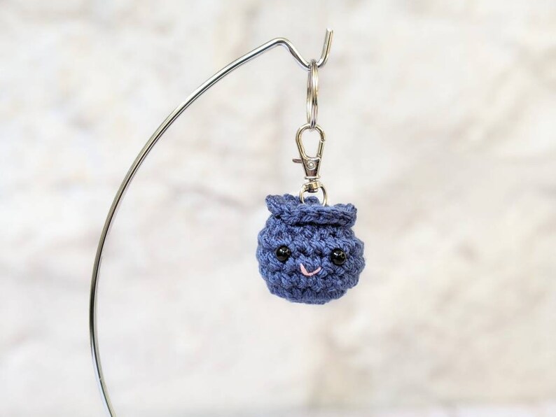 May include: A small, navy blue crocheted keychain shaped like a smiling blueberry. The blueberry has black bead eyes and a pink stitched mouth. It is attached to a silver-colored keychain clip.