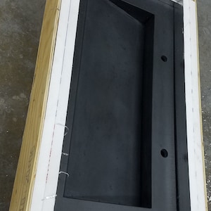 Op de afbeelding: Een groot, zwart, rechthoekig object met een verzonken ontwerp, ingesloten in een houten kist. De kist heeft witte en zilveren isolatie en is vastgezet met witte tape. Het object heeft twee ronde gaten.