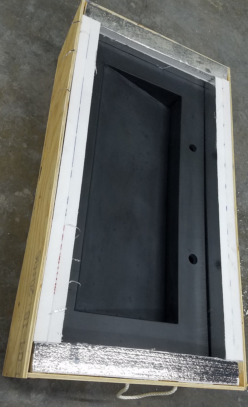Op de afbeelding: Een groot, zwart, rechthoekig object met een verzonken ontwerp, ingesloten in een houten kist. De kist heeft witte en zilveren isolatie en is vastgezet met witte tape. Het object heeft twee ronde gaten.
