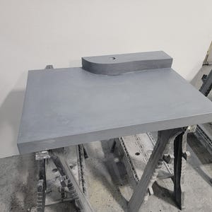 Op de afbeelding: Een grijze rechthoekige aanrecht met een verhoogd, gebogen gedeelte en een cirkelvormig gat. Het aanrecht wordt ondersteund door twee zwarte zaagbokken. Het oppervlak heeft een gladde, matte afwerking.