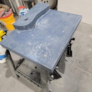 Op de afbeelding: Een rechthoekige, blauwgrijze werktafel met een gebogen verlenging en een gat. De tafel wordt ondersteund door een zwarte kunststof zaagbok. Het oppervlak heeft een textuur met lichtgekleurde spikkels.