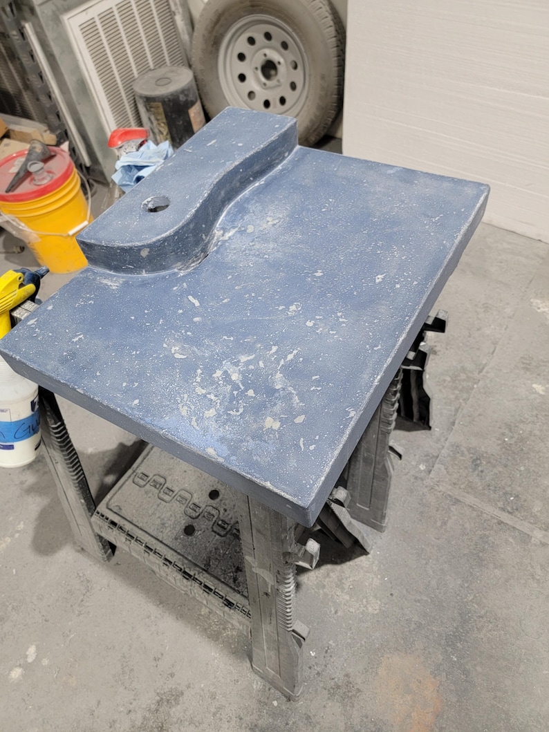 Op de afbeelding: Een rechthoekige, blauwgrijze werktafel met een gebogen verlenging en een gat. De tafel wordt ondersteund door een zwarte kunststof zaagbok. Het oppervlak heeft een textuur met lichtgekleurde spikkels.