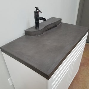Op de afbeelding: Een moderne badkamermeubel met een donkergrijze betonnen aanrechtblad en een zwarte kraan. De wastafel heeft witte kastdeuren met verticale groeven. De kraan heeft een unieke, gebogen uitloop.