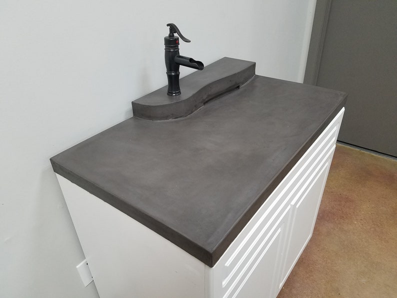 Op de afbeelding: Een moderne badkamermeubel met een donkergrijze betonnen aanrechtblad en een zwarte kraan. De wastafel heeft witte kastdeuren met verticale groeven. De kraan heeft een unieke, gebogen uitloop.