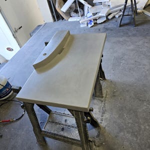 Op de afbeelding: Een lichtgrijze betonnen aanrechtblad met een gebogen achterwand en een rond gat. Het aanrechtblad staat op een donkergrijze metalen zaagbok in een werkplaats. Het aanrechtblad is ongeveer 122 cm lang.