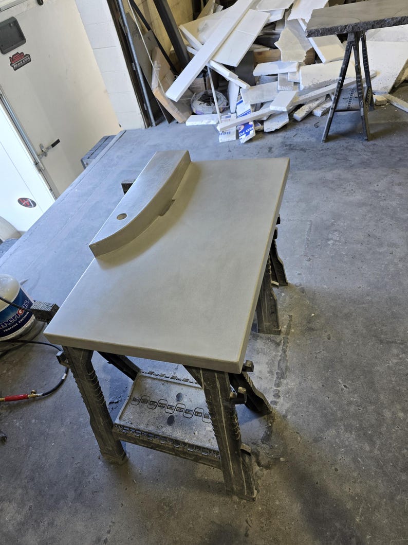 Op de afbeelding: Een lichtgrijze betonnen aanrechtblad met een gebogen achterwand en een rond gat. Het aanrechtblad staat op een donkergrijze metalen zaagbok in een werkplaats. Het aanrechtblad is ongeveer 122 cm lang.