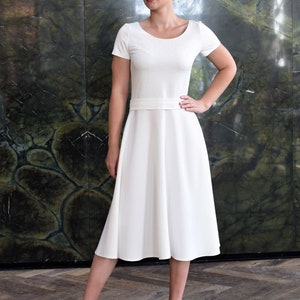 May include: A white dress with a fitted bodice and a full skirt. The dress has short sleeves and a scoop neckline. The model is wearing white heels.