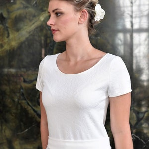 May include: A woman wearing a white dress with a scoop neck and short sleeves. The dress has a textured fabric and a fitted bodice. The skirt is a-line and falls to just above the knees. The woman has blonde hair and is wearing a white flower in her hair.