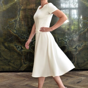 May include: A woman wearing a white dress with a simple design and a white belt. She is standing on a wooden floor and has her hair pulled back. She is wearing white heels.