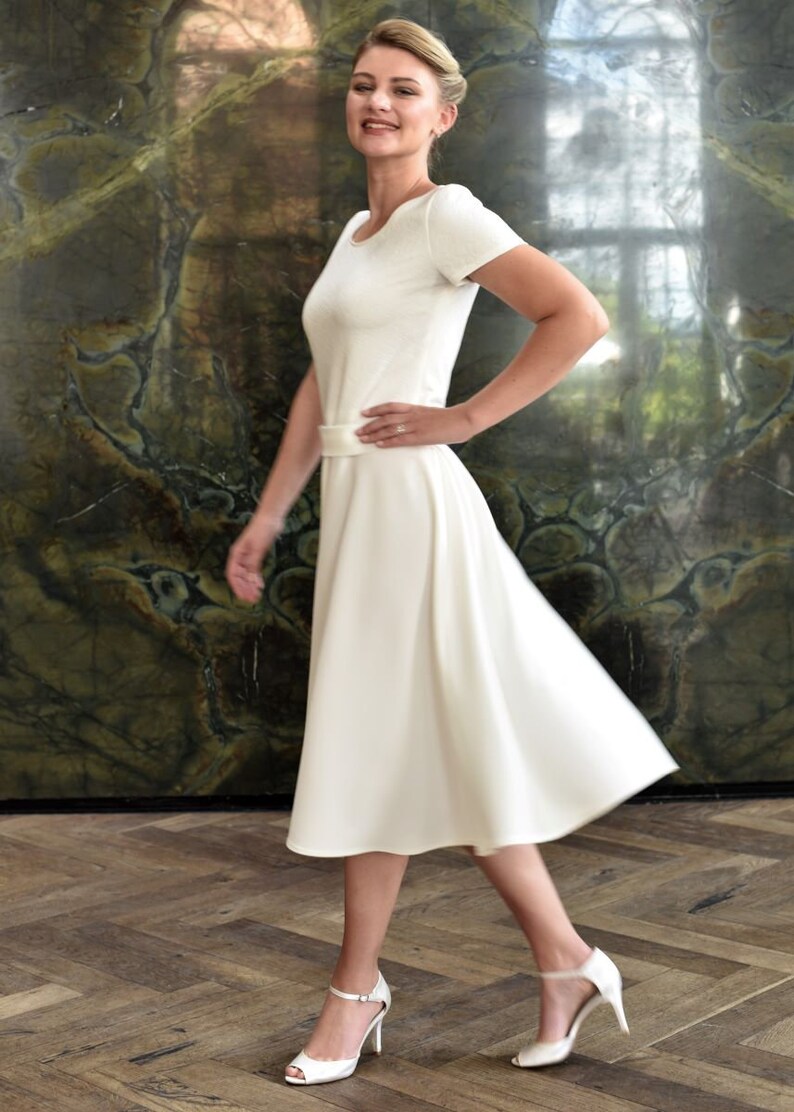 May include: A woman wearing a white dress with a simple design and a white belt. She is standing on a wooden floor and has her hair pulled back. She is wearing white heels.