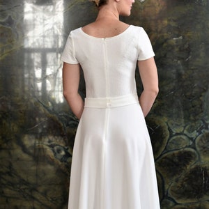 May include: A white dress with a scoop neckline and short sleeves. The dress has a fitted bodice and a full skirt. The dress is cinched at the waist with a belt and has a zipper closure in the back.