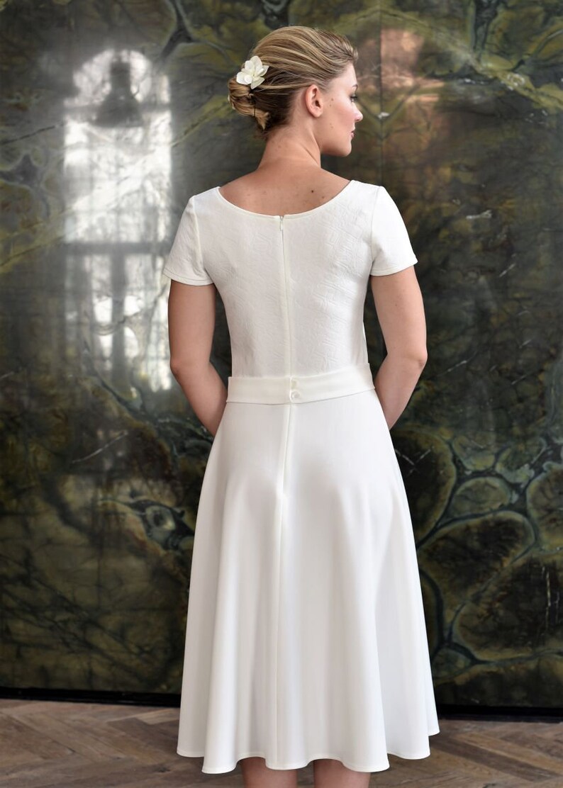 May include: A white dress with a scoop neckline and short sleeves. The dress has a fitted bodice and a full skirt. The dress is cinched at the waist with a belt and has a zipper closure in the back.