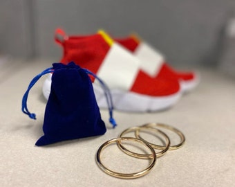 Può includere: Tre anelli d'oro impilati uno sopra l'altro. Un sacchetto di velluto blu si trova su una superficie bianca. Una scarpa rossa e bianca è sfocata sullo sfondo.