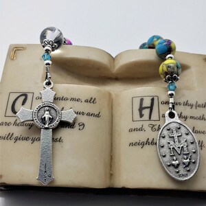 May include: A silver rosary with blue and yellow beads and a silver charm with the Virgin Mary. The rosary is resting on a book with the text "into me, all are heavy and I will give you rest." and "onur thy fath Why moth and, Th neighb love".