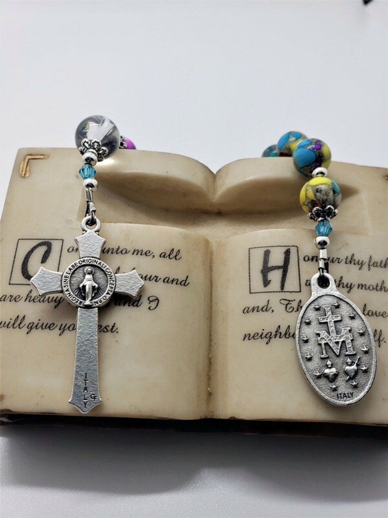 May include: A silver rosary with blue and yellow beads and a silver charm with the Virgin Mary. The rosary is resting on a book with the text "into me, all are heavy and I will give you rest." and "onur thy fath Why moth and, Th neighb love".