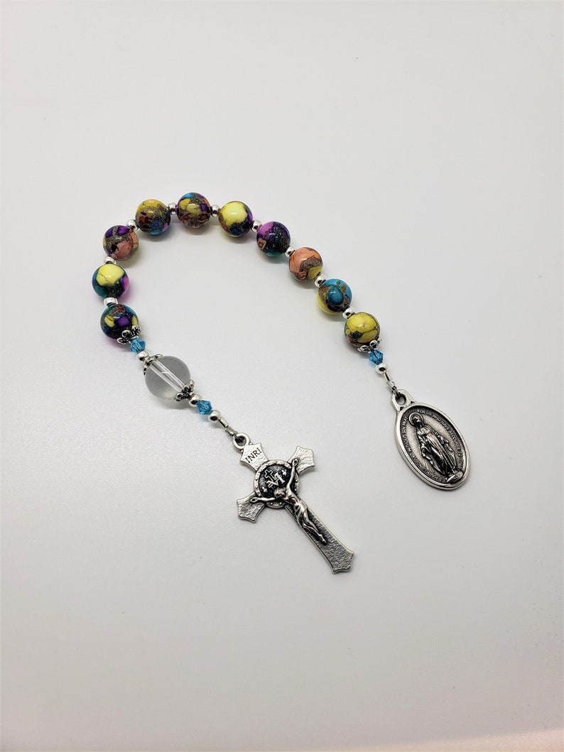 May include: A colorful beaded rosary with a silver crucifix and a silver oval pendant featuring the image of the Virgin Mary. The beads are multi-colored with shades of yellow, orange, purple, and blue.