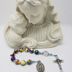 May include: A colorful rosary with a clear crystal bead, a silver charm of the Virgin Mary, and a silver crucifix with the inscription "INRI". The rosary is displayed in front of a white statue of the Virgin Mary and child.