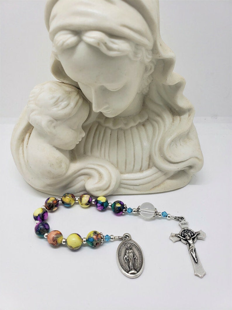 May include: A colorful rosary with a clear crystal bead, a silver charm of the Virgin Mary, and a silver crucifix with the inscription "INRI". The rosary is displayed in front of a white statue of the Virgin Mary and child.
