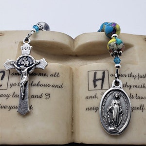 May include: Two silver religious rosaries with blue and yellow beads. One rosary features a silver crucifix with a figure of Jesus Christ. The other rosary features a silver oval pendant with an image of the Virgin Mary. The rosaries are displayed on a book with text.