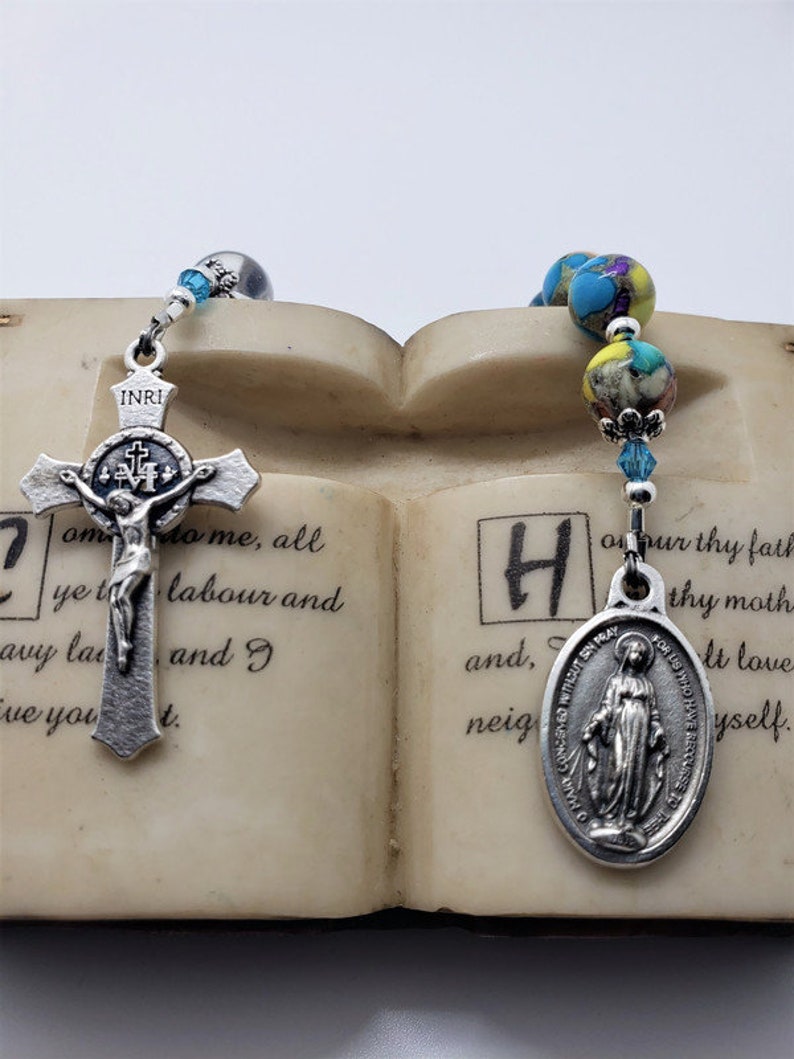 May include: Two silver religious rosaries with blue and yellow beads. One rosary features a silver crucifix with a figure of Jesus Christ. The other rosary features a silver oval pendant with an image of the Virgin Mary. The rosaries are displayed on a book with text.