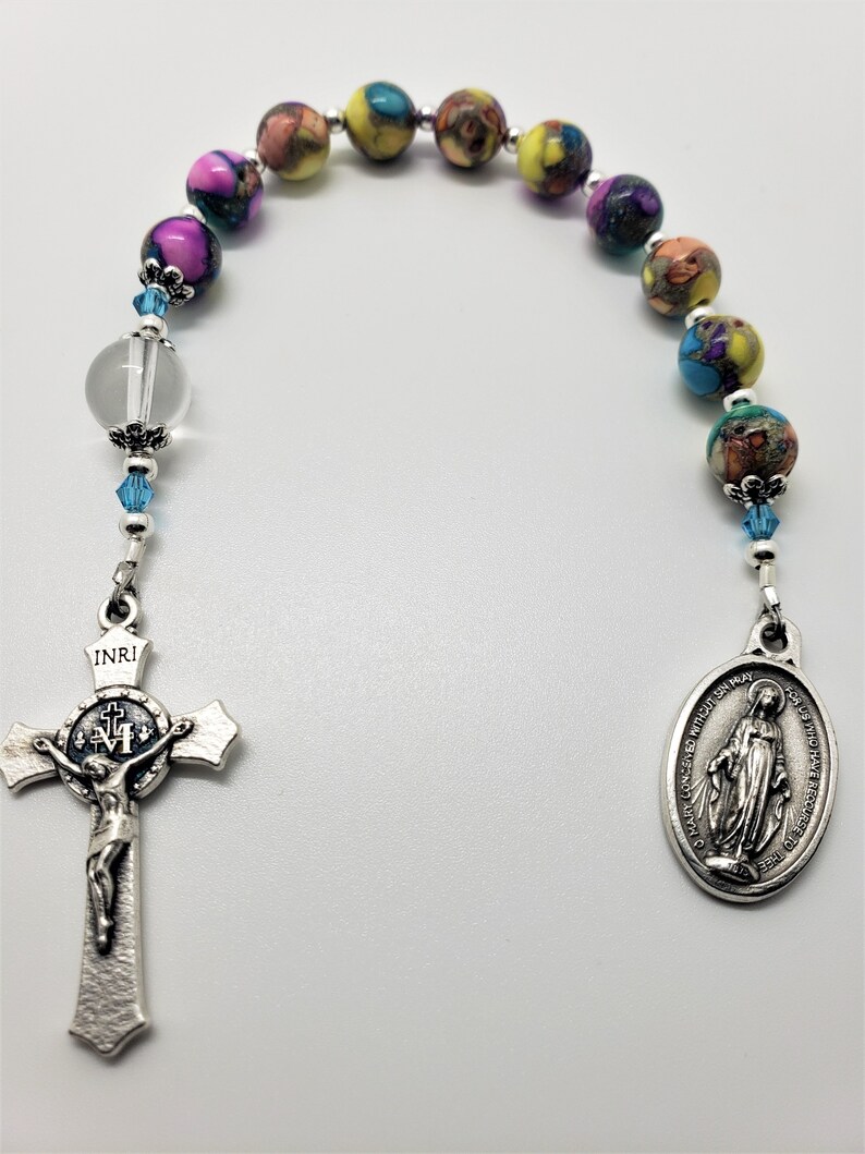 May include: A colorful rosary with a silver crucifix and a silver medal of the Virgin Mary. The beads are made of glass and have a variety of colors, including yellow, blue, pink, and green. The crucifix has the inscription "INRI" on the top and a depiction of Jesus Christ on the cross. The medal has the inscription "O Mary conceived without sin, pray for us who have recourse to thee" on the back.