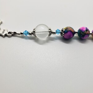 May include: A close-up of a beaded bracelet with a clear crystal bead, blue crystal beads, and pink and green patterned beads.