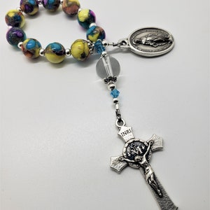 May include: A colorful beaded rosary with a silver crucifix and a silver oval pendant featuring the Virgin Mary. The beads are yellow, blue, and purple with a marbled pattern.