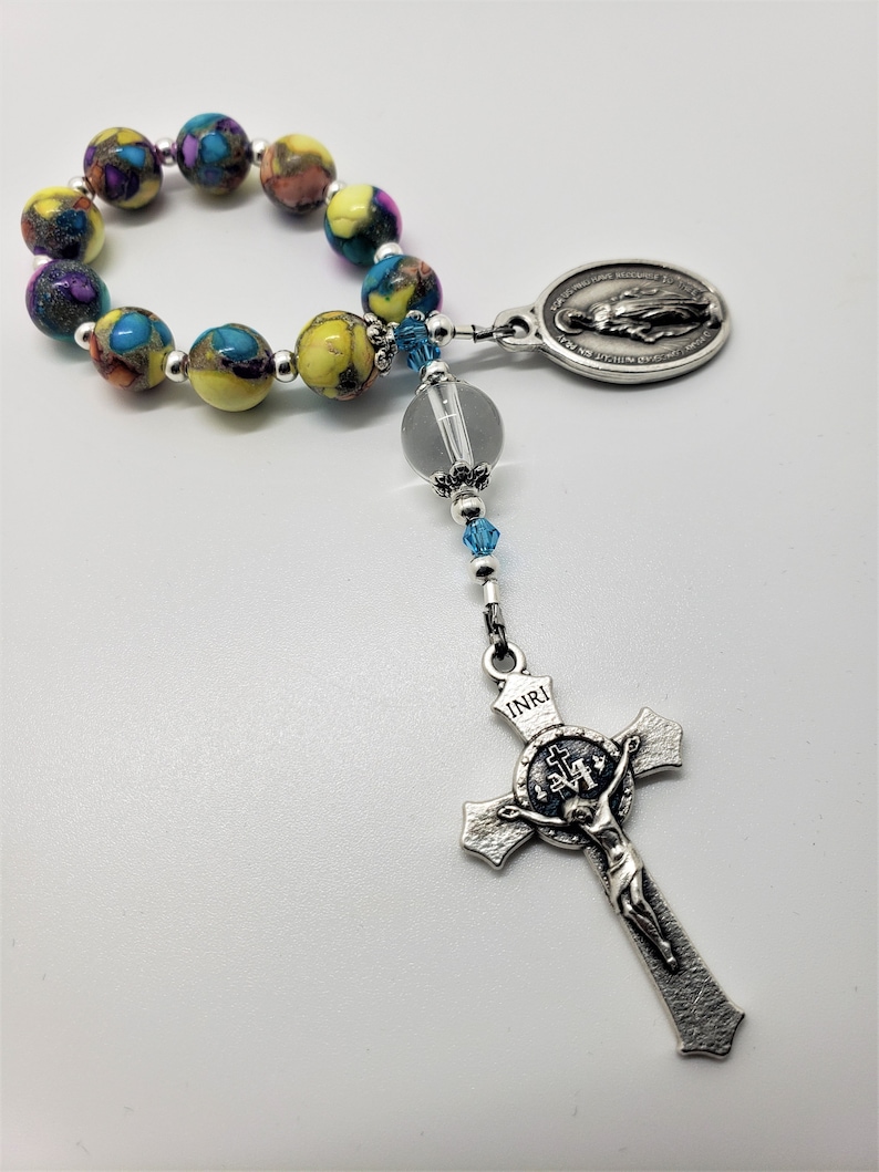 May include: A colorful beaded rosary with a silver crucifix and a silver oval pendant featuring the Virgin Mary. The beads are yellow, blue, and purple with a marbled pattern.