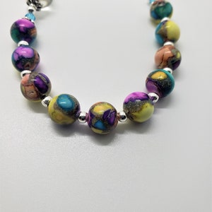 May include: A colorful beaded bracelet with a silver charm featuring a religious image. The beads are multi-colored and have a speckled pattern.