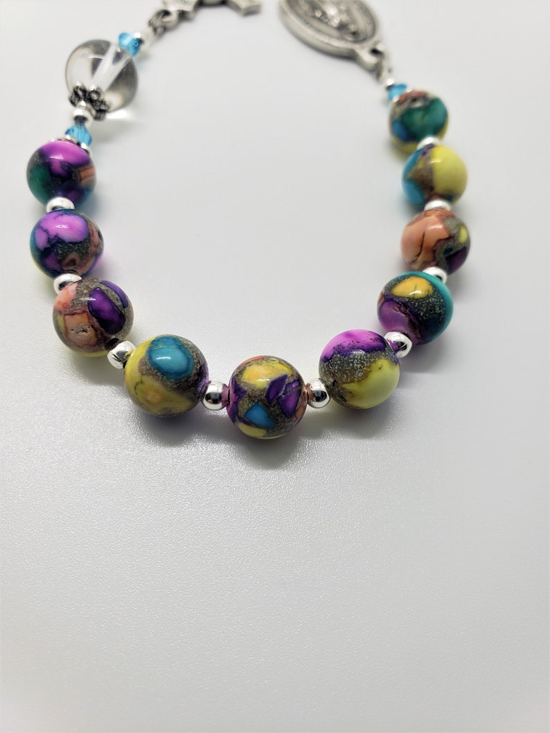 May include: A colorful beaded bracelet with a silver charm featuring a religious image. The beads are multi-colored and have a speckled pattern.