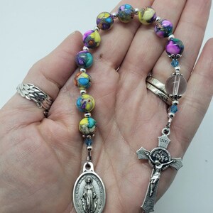 May include: A colorful beaded rosary with a silver cross and a silver oval charm featuring the image of the Virgin Mary. The beads are made of a multi-colored stone with a speckled pattern.