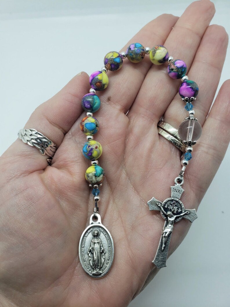 May include: A colorful beaded rosary with a silver cross and a silver oval charm featuring the image of the Virgin Mary. The beads are made of a multi-colored stone with a speckled pattern.
