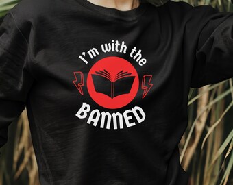 May include: Black long-sleeved sweatshirt with the text "I'm with the Banned" in white, centered on the chest. The text is above and below a red circle with a book graphic and lightning bolts. The sweatshirt is a casual style.
