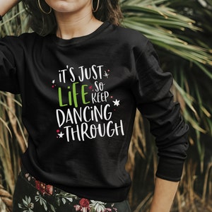 以下が含まれることがあります： 白い文字で「It's just life so keep dancing through」と書かれた黒い長袖スウェットシャツ。テキストは赤と白の小さな星で飾られています。