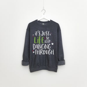 以下が含まれることがあります： 緑と赤のアクセントが付いた白い文字で「it's just life so keep dancing through」と書かれた濃いグレーのスウェットシャツです。