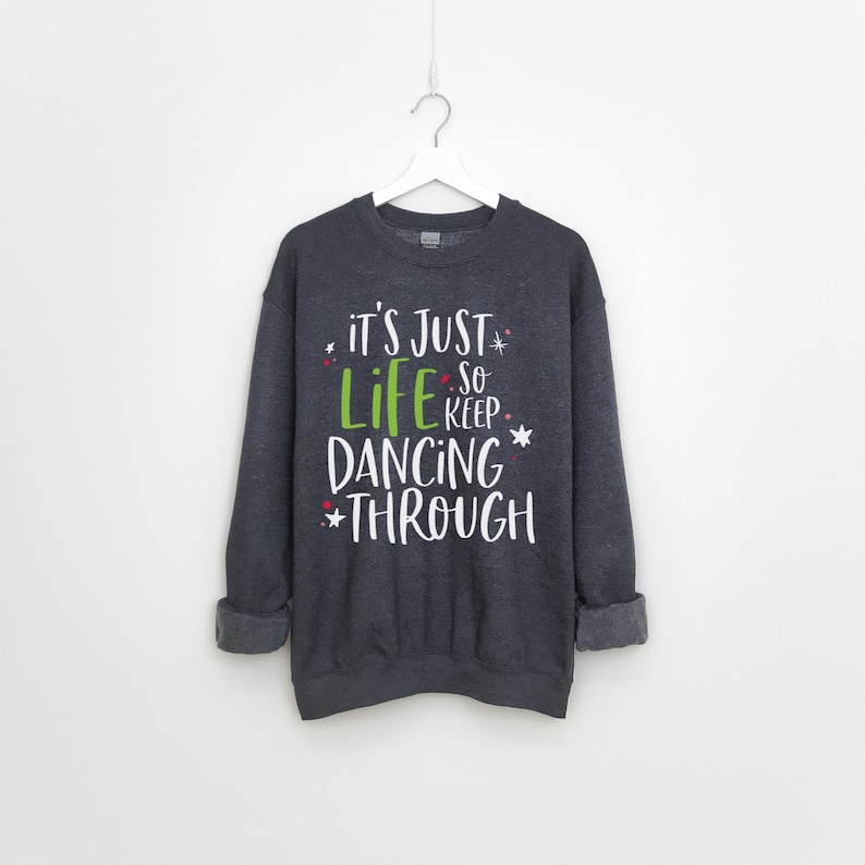 以下が含まれることがあります： 緑と赤のアクセントが付いた白い文字で「it's just life so keep dancing through」と書かれた濃いグレーのスウェットシャツです。