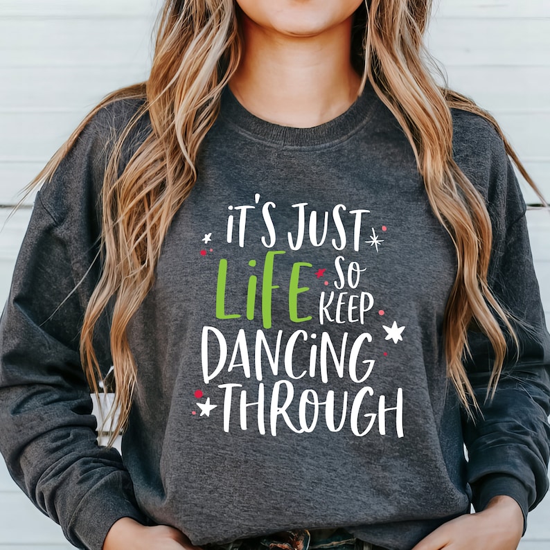 以下が含まれることがあります： グレーのトレーナーに「It's just life so keep dancing through」という白い文字と緑の文字で書かれたテキストが入っています。