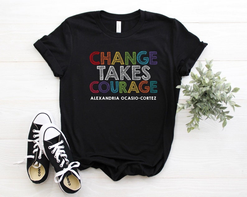 May include: Black t-shirt with the text "CHANGE TAKES COURAGE" in rainbow colors and the text "ALEXANDRIA OCASIO-CORTEZ" in white below.