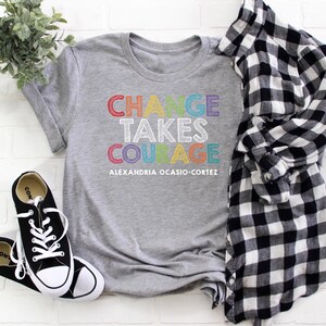 May include: Gray t-shirt with the text "CHANGE TAKES COURAGE" in rainbow colors and the text "ALEXANDRIA OCASIO-CORTEZ" in black below.