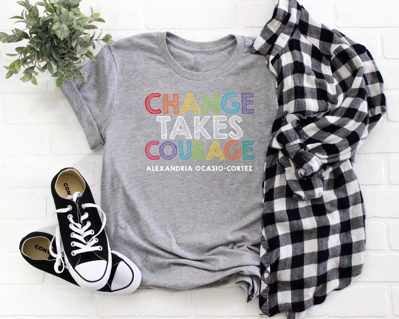 May include: Gray t-shirt with the text "CHANGE TAKES COURAGE" in rainbow colors and the text "ALEXANDRIA OCASIO-CORTEZ" in black below.
