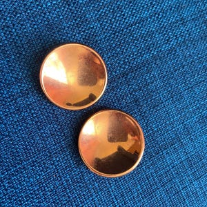 May include: Two circular, metallic copper-colored buttons with a smooth, reflective, and slightly concave surface. These buttons feature a polished finish that catches the light, creating a bright sheen across their rounded, bowl-like shape.